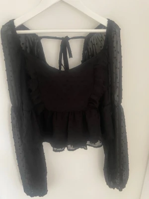 Svart blus med volang och knyt - Snygg svart blus med transparenta, prickiga långärmar och volangdetaljer. Toppen har knyt i ryggen och lätt puffiga ärmar för en trendig look. Perfekt att styla med jeans eller kjol för en cool outfit. 