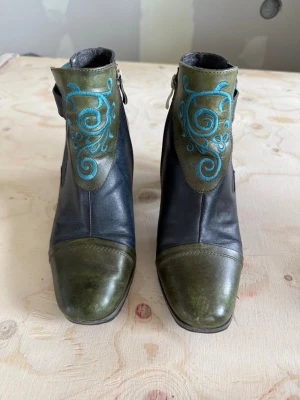 Unika stövletter med brodyr - Snygga stövletter i svart och olivgrönt läder med turkos broderi framtill. Skorna har rundad tå, träklack och dekorativ knappdetalj på sidan. Dragkedja på insidan för enkel påtagning. Perfekt för dig som vill sticka ut med din stil.