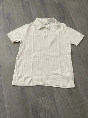 Vit ribbad polotopp från Kappahl - Klassisk vit polotopp från Kappahl med ribbad struktur och korta ärmar. Toppen har krage och knappar framtill för en stilren look. Perfekt till jeans eller kjol för en clean och enkel outfit.
