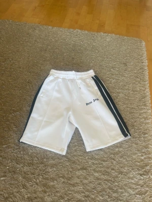 Vita Palm Angels shorts med svarta ränder - Palm Angels vita shorts med svarta sidoremsor och elastisk midja med snörning. Klassisk sportig look med logotyp på vänster ben. Perfekta för en avslappnad och trendig stil.