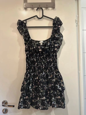 Svart blommig kortklänning med volang - Supersöt svart kortklänning med blommigt mönster och volangdetaljer. Klänningen har breda axelband, smockad midja och volanger upptill och nertill. Perfekt för sommarens alla tillfällen och ger en romantisk vibe.