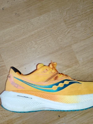 Gula Saucony löparsneakers strlk 42.5 - Säljer ett par snygga gula sneakers från Saucony med blå och orange detaljer. Skorna har en låg profil, mesh-överdel för bra ventilation och tjock, vit sula med PWRRUN+ teknologi för extra komfort. Perfekta för dig som gillar sportig stil och vill sticka ut. Skorna är i mkt bra skick, endast använda ett fåtal gånger.  Nypris unt 2-2500:-