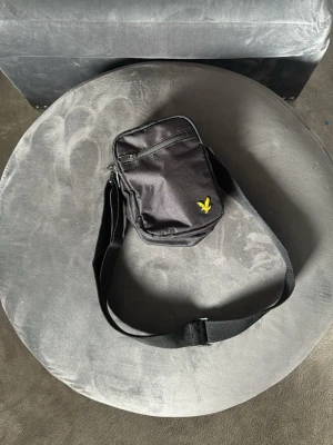 Svart axelväska från Lyle & Scott - Snygg svart axelväska från Lyle & Scott med det klassiska gula örnloggan på framsidan. Väskan är tillverkad i slitstark textil och har justerbar axelrem. Perfekt för att bära det viktigaste och ger en clean streetstyle-vibe.