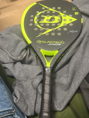 Galactica Junior padelracket - Säljer ett Galactica Junior padelracket i svart och neongult med kolfibermönster. Racketen har perforerade hål och signaturdetalj på framsidan. Perfekt för juniorer som vill ta sitt padelspel till nästa nivå.