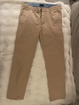 Beiga chinos från GANT - Snygga beiga chinos från GANT med klassisk rak passform och diskreta sidofickor. Tillverkade i mjukt bomullstyg med blåvitrandigt innerfoder i midjan. Perfekta för en clean och stilren look.