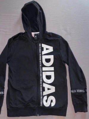 Svart hoodie från Adidas - Svart hoodie från Adidas med stor vit logga längs dragkedjan framtill och textdetaljer på ärmarna. Tröjan har huva. 
