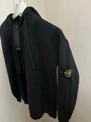 Svart hoodie från Stone Island - Snygg svart hoodie från Stone Island med klassisk patch på ärmen. Hoodien har dragsko i huvan och en stor magficka framtill. Tillverkad i mjukt bomullsmaterial och har ribbade muddar vid ärmslut och nederkant. Perfekt för en avslappnad streetwear-look.