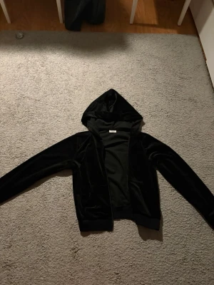 Svart hoodie med huva - Snygg svart hoodie i mjuk velour med dragkedja framtill och huva. Tröjan har långa ärmar och ribbade muddar vid ärmslut och nederkant. Perfekt för en avslappnad och trendig look.