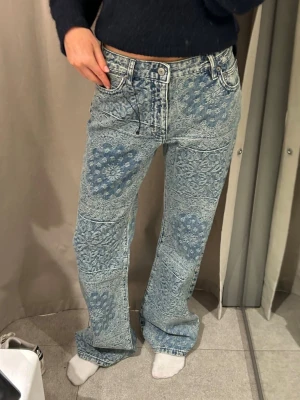 Blå mönstrade raka jeans Zara - Unika blå jeans från Zara med patchwork-liknande blommönster över hela byxan. Modellen är rak med hög midja och klassiska fem fickor. Jeansen har en cool vintagekänsla och är tillverkade i denim med tydliga sömmar och knappgylf.