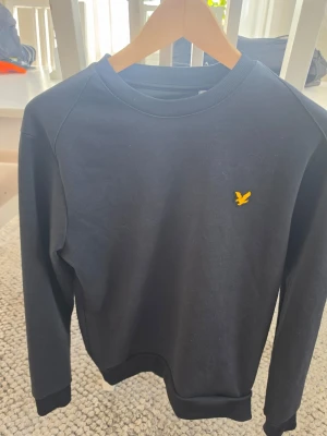 Svart tröja från Lyle & Scott - Klassisk svart tröja från Lyle & Scott med rund halsringning och den ikoniska gula fågeln broderad på bröstet. Tröjan har långa ärmar och ribbade muddar vid ärmslut och nederkant. Perfekt till en enkel och stilren look.