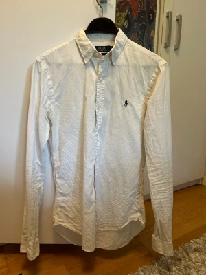 Vit skjorta från Polo Ralph Lauren - Klassisk vit skjorta från Polo Ralph Lauren med broderad logga på bröstet. Skjortan har lång ärm, knappar framtill och en tidlös design som passar till många olika stilar.