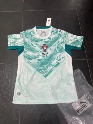 Portugal fotbollströja från Puma - Snygg Portugal fotbollströja från Puma i mintgrönt och vitt med vågigt mönster. Tröjan har korta ärmar, rund hals och Portugal-emblemet på bröstet. Tillverkad i lätt och ventilerande material, perfekt för träning eller match.