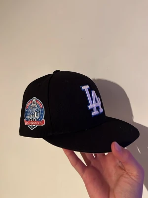 Svart LA Dodgers keps New Era - Säljer en svart fitted keps från New Era med LA Dodgers logga i vitt framtill och en broderad patch på sidan. Baksidan har MLB-loggan. Klassisk rak skärm och snygg streetstil, perfekt för dig som gillar sportiga accessoarer.