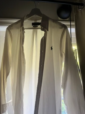 Ralph Lauren - Klassisk vit o blå skjorta från Ralph Lauren med lång ärm och knappar framtill. Skjortan har den ikoniska broderade loggan på bröstet och är tillverkad i bomull för en skön och stilren känsla. 