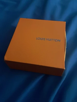 Louis Vuitton ask med tygpåse - Säljer en snygg orange ask från Louis Vuitton med tillhörande beige tygpåse. Perfekt för att förvara accessoarer eller smycken. Asken är i hård kartong och har klassisk Louis Vuitton-logga på locket.