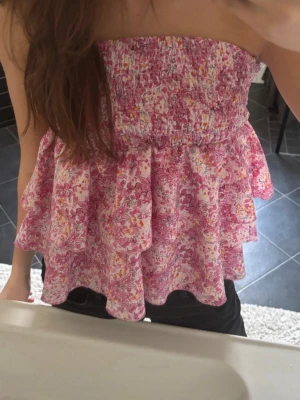 Blommig bandeau topp i rosa toner - Säljer en söt bandeau topp med volanger och blommigt mönster i rosa, vitt. Knappt använt. töjbar topp o formas fint efter dig så passar egentligen S,M och L