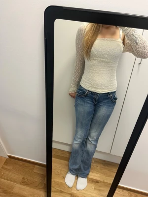Vit spetsblus med långa ärmar - Säljer en vit, figurnära topp i spets med långa ärmar och smala axelband. Toppen har ett vackert blommönster i spetsen och lätt rynkad front. Perfekt för dig som gillar romantisk och trendig stil.