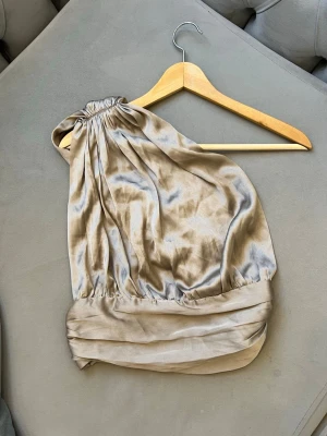 Champagnefärgad oneshoulder topp i satin - Snygg oneshoulder topp i champagnefärgad satin med bred draperad kant nedtill och lätt glansigt tyg. Perfekt för dig som vill ha en unik och trendig look. Toppen har  ärmlös design med fina veckdetaljer. Toppen är helt ny och oanvänd!