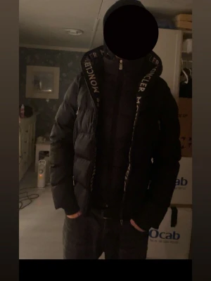 Svart pufferjacka från Moncler - Snygg svart pufferjacka från Moncler med stor huva och logotyp på insidan av kragen. Jackan har en quiltad design och dragkedja framtill. Perfekt för kalla dagar när du vill hålla stilen och värmen.