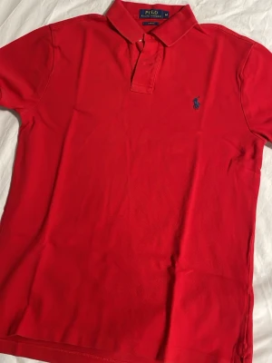 Röd pikétröja från Polo Ralph Lauren - Klassisk röd pikétröja från Polo Ralph Lauren med blå broderad logga på bröstet. Tröjan har krage, korta ärmar och knappar vid halsen. Slim fit-modell i mjuk bomull som passar perfekt till en avslappnad stil.