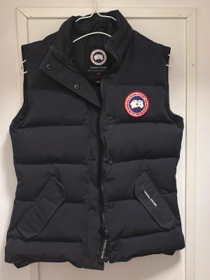 Svart dunväst från Canada Goose - Snygg svart dunväst från Canada Goose med klassisk logga på bröstet och dragkedja framtill. Västen har två snedställda fickor med lock och tryckknappar samt hög krage för extra värme. Perfekt för lager-på-lager och streetstyle.  Skriv för närmare bilder på västen!💗
