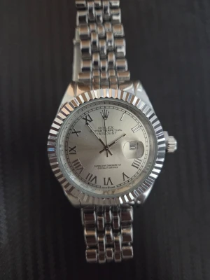 silverfärgad klocka - Klassisk Datejust klocka i silverfärg med romerska siffror på urtavlan och datumvisning. Armbandet är i polerat metall och har en snygg länkdesign. Klockan har räfflad bezel och tydlig logga på spännet.