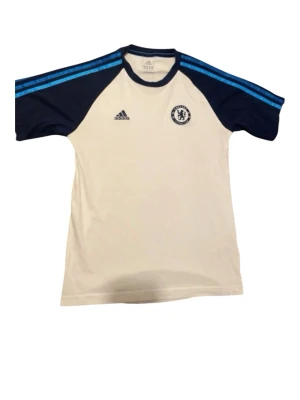 Chelsea FC träningströja Adidas - Snygg vit träningströja från Adidas med Chelsea FC-logga på bröstet. Tröjan har mörkblå ärmar med blå ränder på axlarna och klassisk Adidas-logga. Perfekt för fotbollsträning eller att representera ditt favoritlag. Tillverkad i lätt och funktionellt material. Storlek xs och i ett bra skick
