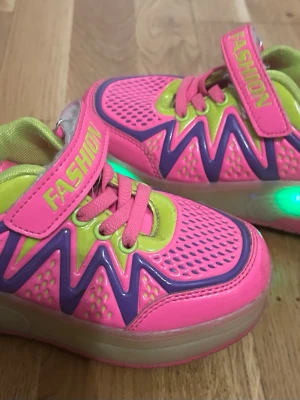Neonrosa sneakers med hjul och LED - Färgglada sneakers i neonrosa med limegröna och lila detaljer, kardborreband och snörning. Skorna har inbyggda hjul i sulan och LED-ljus på sidan. Ovandelen är i syntetmaterial med ventilerande mesh och glansiga partier. Perfekt för dig som vill sticka ut och rulla fram. I strl 32.