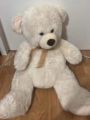 Stor vit nallebjörn med rosett - Mysig och stor nallebjörn i mjuk vit plysch med beige nos och öron. Nallen har en beige rosett runt halsen och svarta ögon. Perfekt som dekoration eller present till någon speciell. 100cm lång