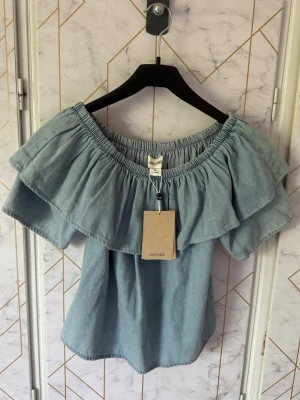 Ljusblå offshoulder topp från Monki - Trendig ljusblå offshoulder topp från Monki. Toppen har bred volang runt hela axelpartiet och en avslappnad passform. Storlek Xs men passar även S