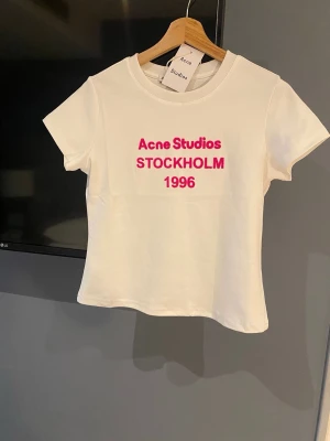 Vit Acne Studios t-shirt - Vit t-shirt från Acne Studios med rosa text 'Acne Studios STOCKHOLM 1996' på bröstet. Klassisk rund halsringning och korta ärmar. Perfekt för en clean och trendig look. Helt ny!