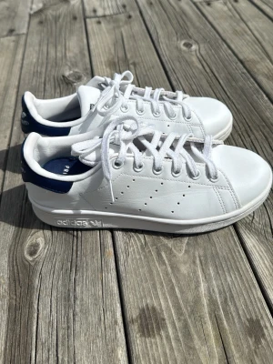 Adidas Stan Smith vita sneakers - Klassiska Adidas Stan Smith sneakers i vitt läder med mörkblå detalj på hälen och vita skosnören. Ikonisk låg modell med perforerade ränder på sidorna och Adidas-logga på sidan och plösen. Perfekta för en clean och stilren look, de är lite slitna men de går att fixa om man vill.