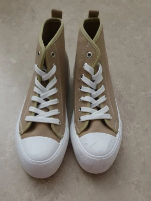 Sneakers - Snygga beige high top sneakers i canvas med vita gummisulor och tåhättor. Skorna har vita skosnören och metallöljetter för en klassisk look. Perfekta för dig som gillar enkel och stilren design.                               Jag testade skorna på butiken och den var i sånt skick men ville köpa ändå då det var i bra pris. Så ordinarie pris var 80kr och nypris 50kr. Så har inte använt den.
