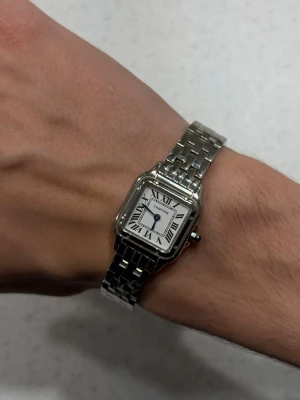 Silverklocka - Klocka i silverfärgat stål med fyrkantig boett och klassisk länkarmband. Vit urtavla med svarta romerska siffror och blå visare ger en tidlös och elegant look. Perfekt accessoar för dig som gillar lyxig och ikonisk design.