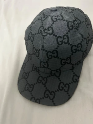 Mörkgrå keps från Gucci med GG-mönster - Snygg mörkgrå keps från Gucci med det klassiska GG-monogrammet över hela kepsen. Tillverkad i ett rutmönstrat tyg som ger en modern känsla. Böjd skärm och justerbar passform bak. Perfekt för dig som vill ha en exklusiv accessoar.