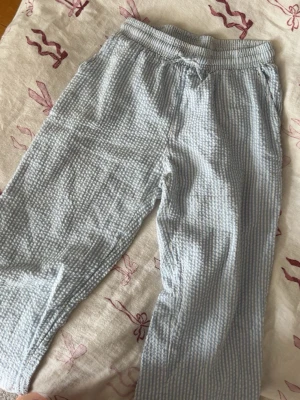 Randiga pyjamasbyxor i bomull - Mysiga pyjamasbyxor med ljusblå och vita ränder i ett luftigt, krinklat bomullstyg. Byxorna har resår i midjan och dragsko för extra komfort. Perfekta för slappa dagar hemma eller en skön natts sömn.