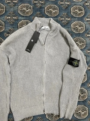Ljusgrå ziptröja från Stone Island - Säljer en ljusgrå stickad ziptröja från Stone Island med hög krage och ribbade muddar. Tröjan har en ikonisk svart patch med gul och grön logga på ärmen. Perfekt för dig som gillar streetwear och snygga detaljer.