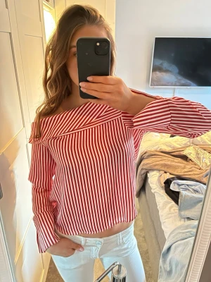Rödvit randig offshoulder blus - Trendig blus med röda och vita vertikala ränder och offshoulder-design. Blusen har långa ärmar och en bred krage som ger en cool och avslappnad look.  Endast använd en gång 