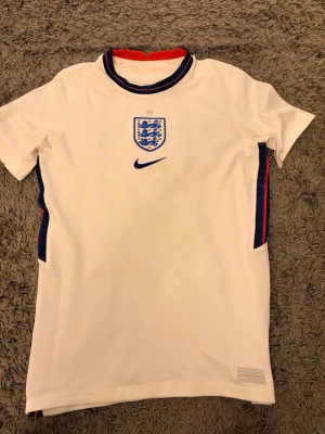 England Nike vit matchtröja - Säljer en vit England matchtröja från Nike med blått och rött mönster på sidorna och kragen. Tröjan har England-emblemet och Nike-loggan på bröstet. Tillverkad i ett lätt och ventilerande material, perfekt för fotboll eller träning.