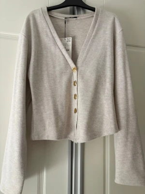 Beige kofta med guldfärgade knappar - Stilren beige kofta med v-ringning och fyra guldfärgade knappar framtill. Koftan har långa, något vida ärmar och är tillverkad i ett mjukt, ribbat material. Perfekt att slänga över en topp för en avslappnad look. Helt oanvänd med prislapp kvar.