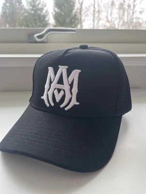 Svart keps från Amiri med logga - Helt oanvänd Snygg svart keps från Amiri med vit broderad logga framtill. Klassisk trucker-modell med mesh på sidorna och justerbar baksida. Tillverkad i bomull och elastan för skön passform. Perfekt accessoar för en trendig streetwear-look.