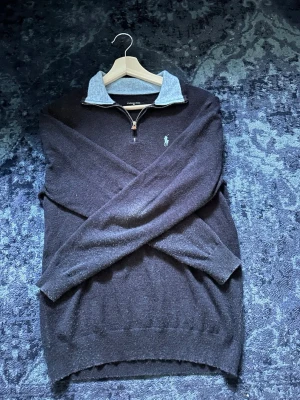 Mörkblå kashmir tröja från Polo Ralph Lauren - Mörkblå stickad tröja från Polo Ralph Lauren med halv dragkedja och ljusblå krage i kontrastmaterial. Klassisk broderad logga i ljusblått på bröstet. Långärmad modell med ribbade muddar och nederkant. Perfekt för en stilren look.