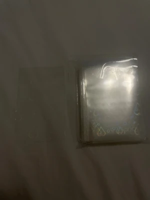 Genomskinliga kort sleeves med hjärtan - Säljer ett paket genomskinliga plast sleeves för samlarkort, dekorerade med holografiska hjärtan och små cirklar längst ner. Perfekt för att skydda dina favoritkort. Materialet är plast och de är genomskinliga med skimrande detaljer.