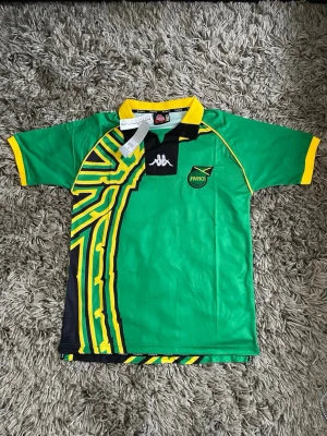 Jamaica fotbollströja från Kappa - Snygg grön Jamaica fotbollströja från Kappa med svart och gul grafisk design på sidan, broderad logga och gul krage. Perfekt för dig som vill sticka ut på planen eller visa ditt stöd för laget. Materialet är lätt och andas, med klassisk sportig passform.