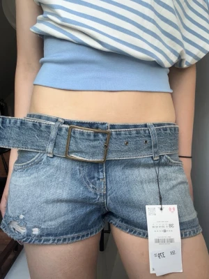 Lågmidjade jeansshorts med bälte - Säljer ett par snygga blå denimshorts med låg midja och tillhörande brett bälte med metallspänne. Midjemåttet är 41 cm sida till sida och passar mig som brukar bära storlek 36-38! Dessa är enbart testade och prislappen finns kvar.