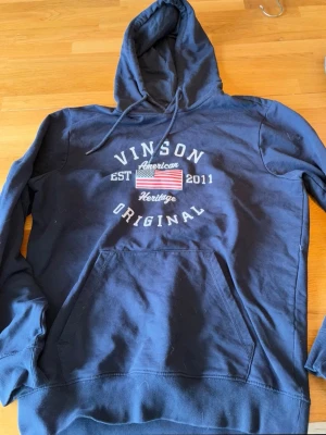 Mörkblå hoodie från Vinson - Mörkblå hoodie från Vinson med tryck framtill som visar amerikansk flagga och texten 'Vinson Original'. Tröjan har huva med dragsko och en stor magficka. Tillverkad i mjukt bomullsmaterial, perfekt för chill dagar.