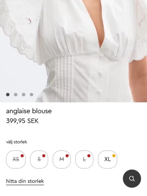 Vit anglaise blus med broderi - Superfin vit blus i anglaise-stil med broderade detaljer och djup v-ringning. Blusen har korta, vida ärmar med vågig kant och markerad midja med sömmar. Perfekt att styla med jeans eller kjol för en trendig look.