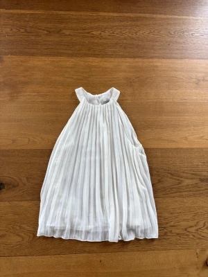 Vit plisserad klänning från H&M - Säljer en elegant vit plisserad klänning från H&M. Klänningen är ärmlös med hög hals och har ett luftigt, flowy fall. Perfekt för dig som gillar en stilren look med fina detaljer. Dragkedja i sidan för enkel påklädning.