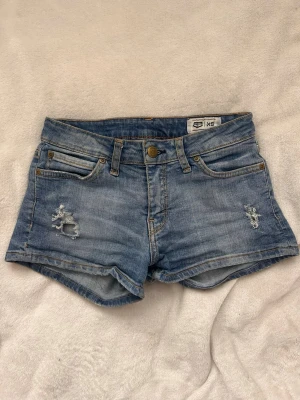 Blå denimshorts från Lee - Säljer ett par klassiska blå denimshorts från Lee med slitna detaljer framtill och femficksmodell. Shortsen har låg midja, knappgylf och en grön Lee-etikett bak. Perfekta för varma dagar och enkel att matcha med allt.