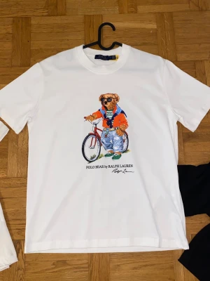 Vit Polo Bear t-shirt från Ralph Lauren - Vit t-shirt från Ralph Lauren med ikoniskt Polo Bear-tryck framtill där björnen står bredvid en cykel i färgglada kläder. Klassisk rund hals och korta ärmar. Perfekt för dig som gillar streetstyle och vill ha något lekfullt i garderoben.